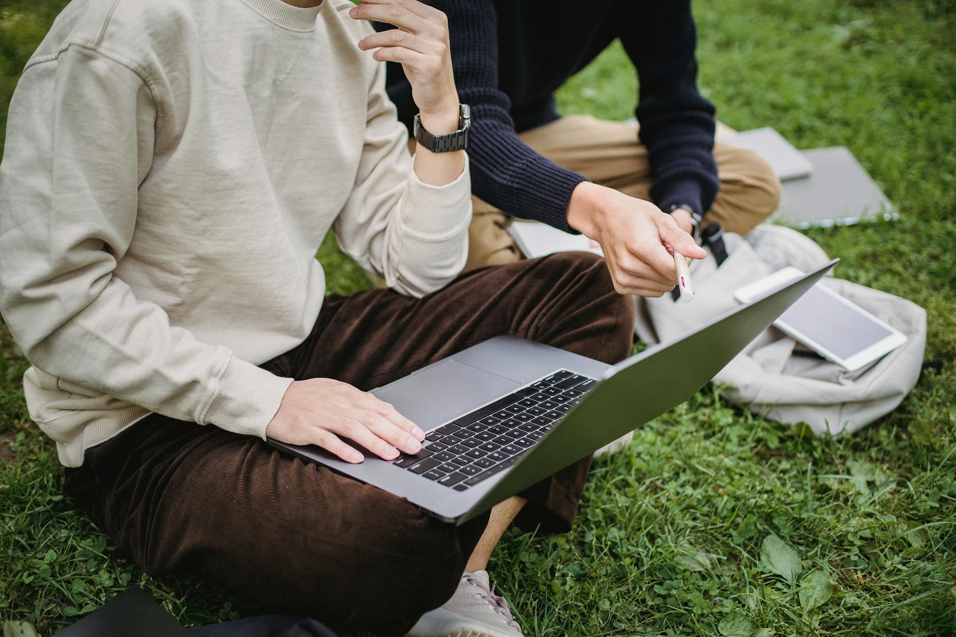 Crop anonymous male student in smart casual wear browsing netbook and discussing university project while sitting on grass in lush garden